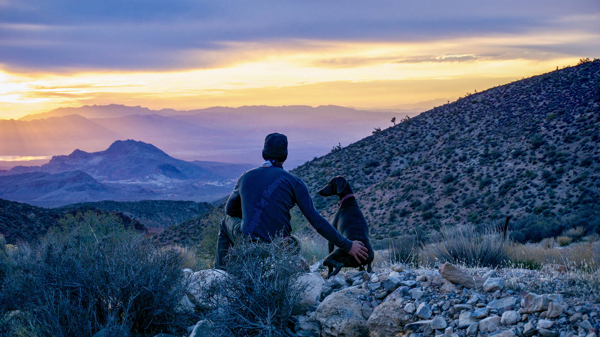 Man and dog overlook a rugged desert valley at sunset, wearing Rugged by Nature® long sleeve thermal shirt – Made in USA outdoor gear.